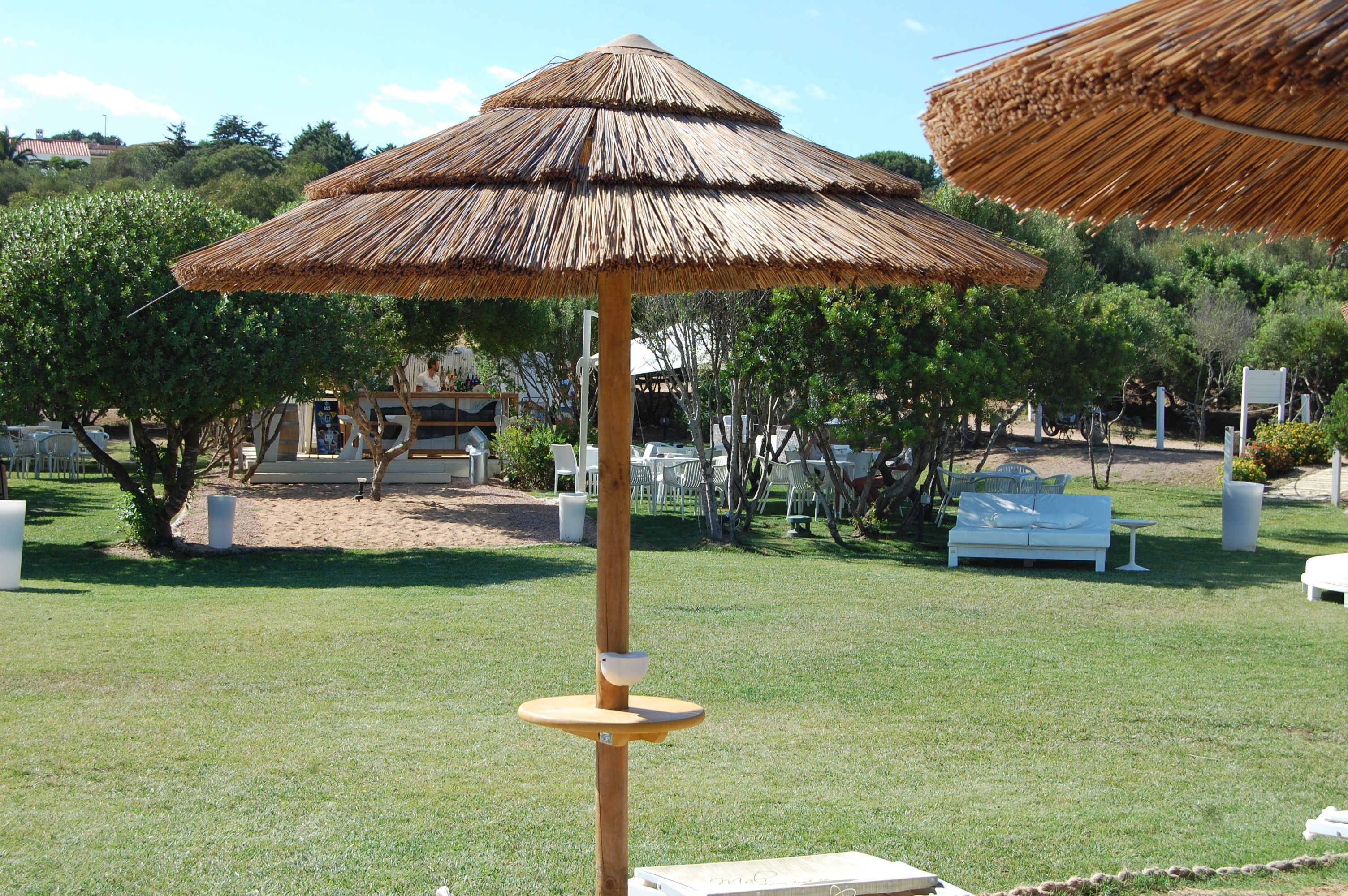 A thatched umbrella structure in a grassy outdoor area with white furniture and trees in the background.