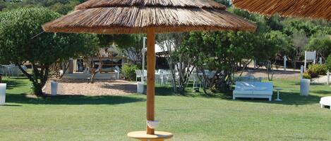 A thatched umbrella structure in a grassy outdoor area with white furniture and trees in the background.