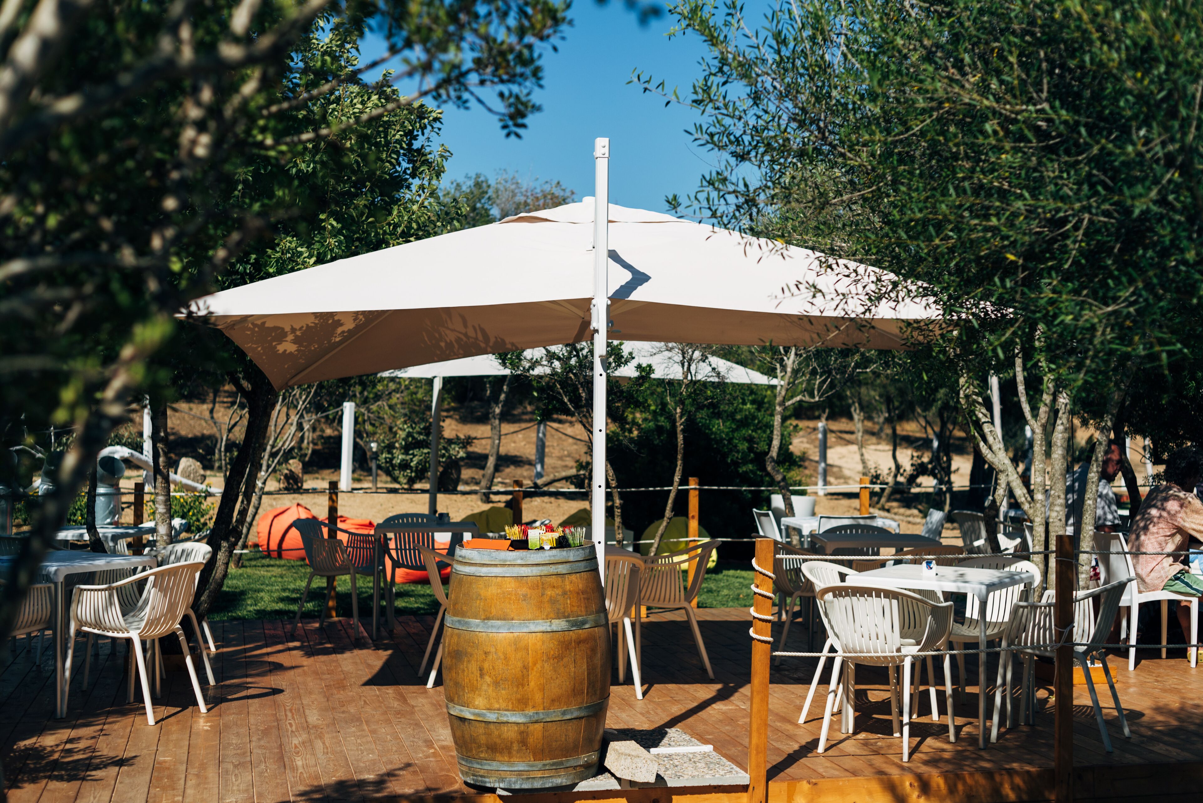 An outdoor dining area with tables and chairs, a wooden barrel, and a large umbrella providing shade.