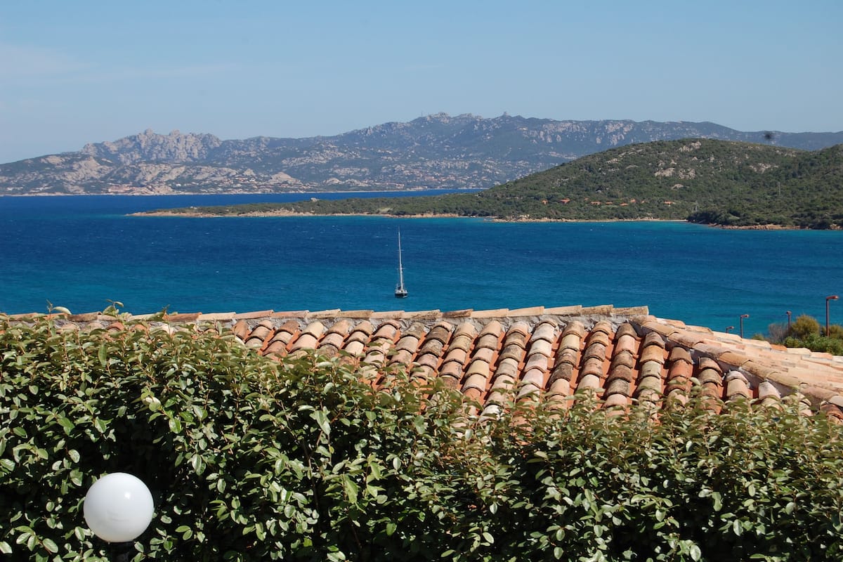 A coastal view with a tiled roof, a sailboat, and distant mountains.