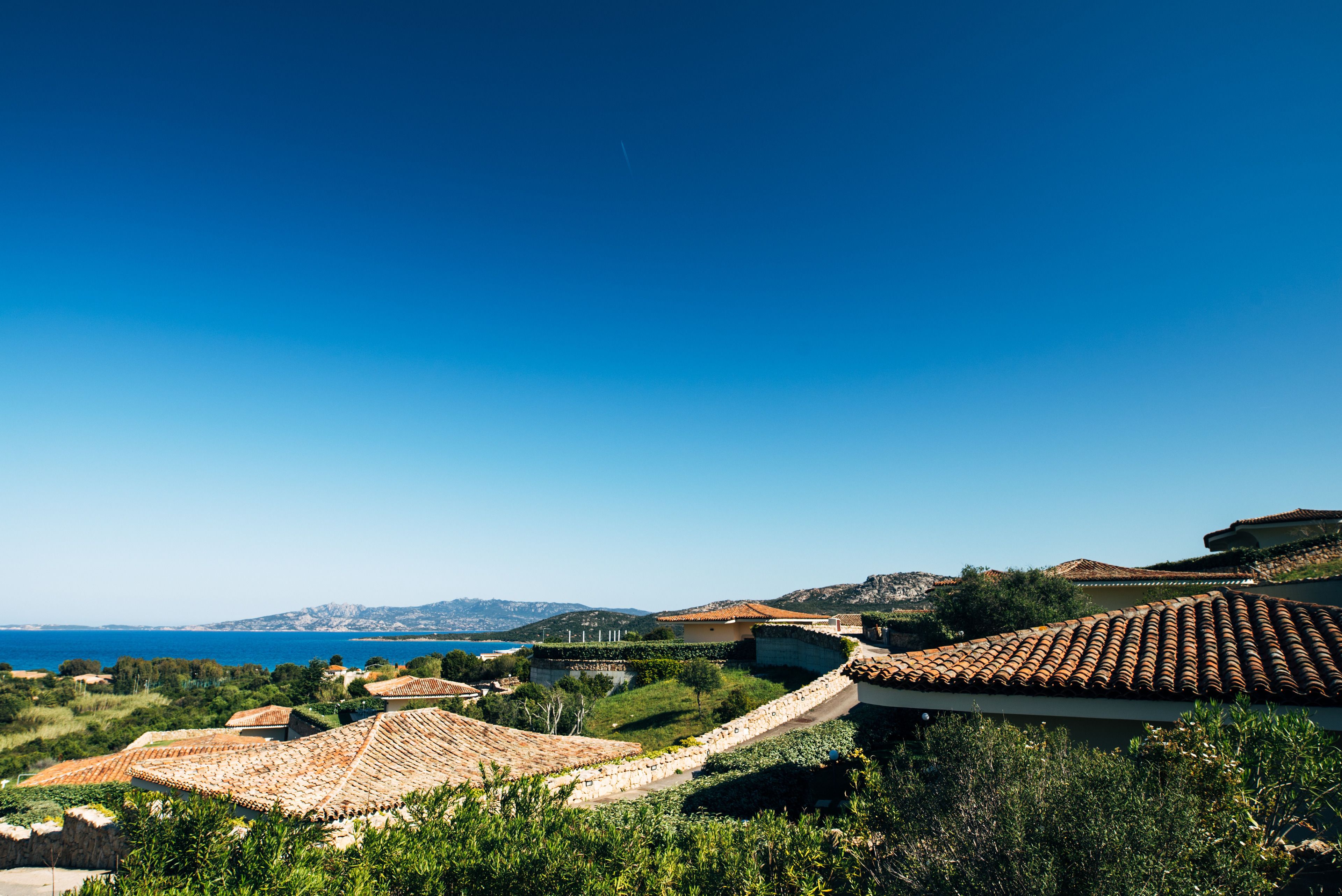 A coastal property with terracotta-tiled roofs, a stone wall, and a view of the sea and distant hills.