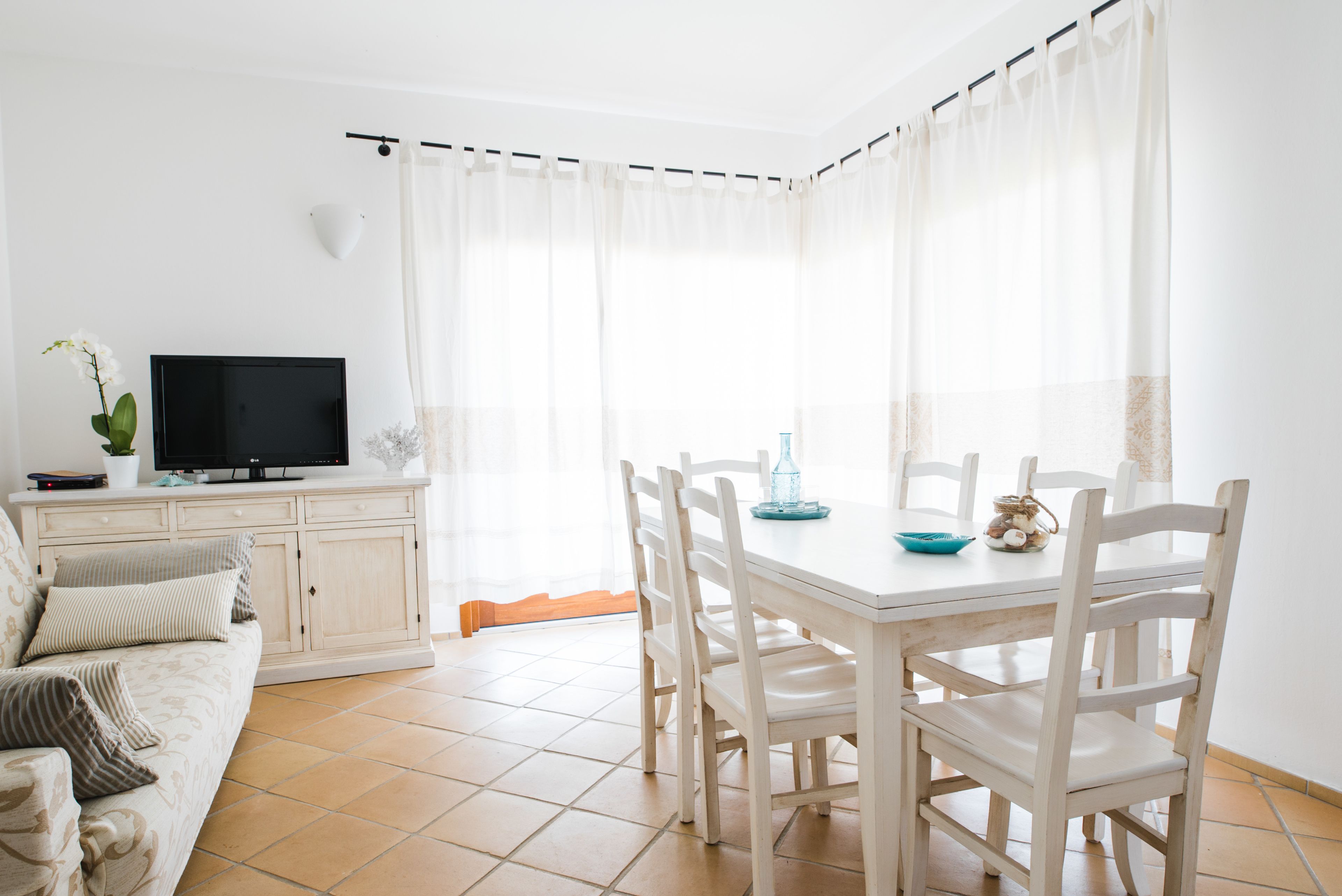 A bright living room with a TV, a sofa, a dining table with chairs, and a large window with curtains.