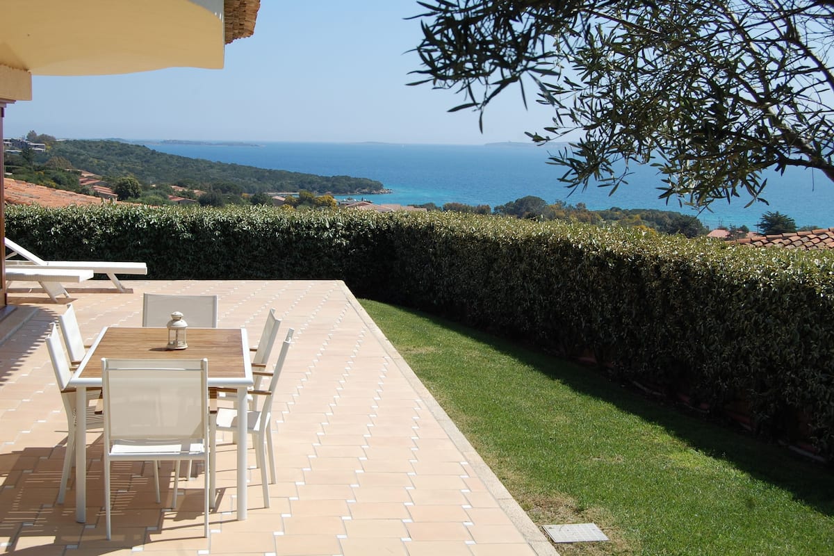 An outdoor patio with a table and chairs, a view of the sea, and a neatly trimmed hedge.