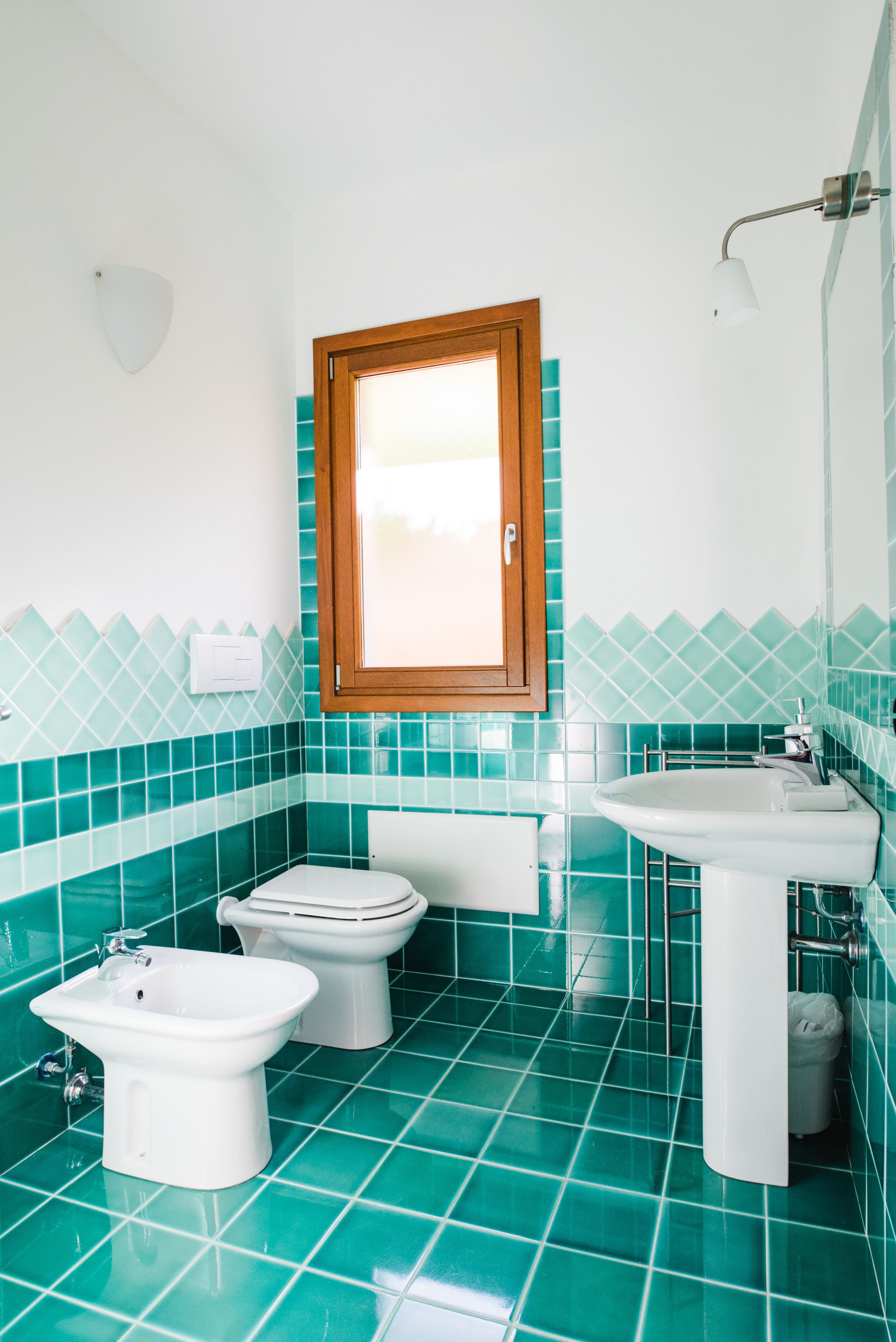 A bathroom with a toilet, bidet, and sink, all in white, with teal tiles on the walls and floor.