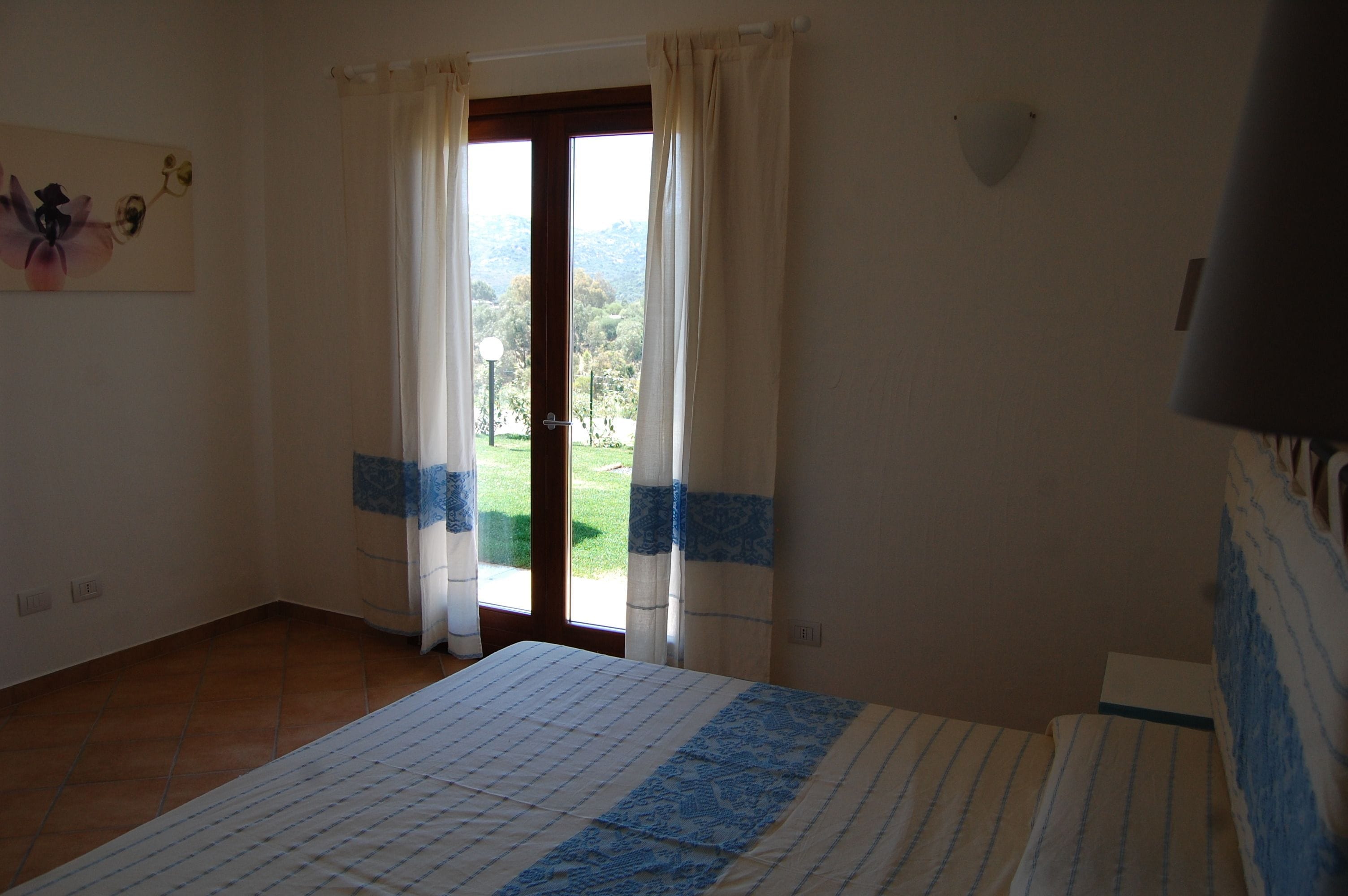Una habitación con cama, una ventana con cortinas y vistas al exterior, además de una lámpara fijada a la pared.