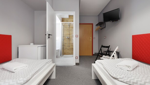 A hotel room with two beds, a shower area, a television mounted on the wall, and a wooden door. - Moon Hostel Kraków (Kraków)