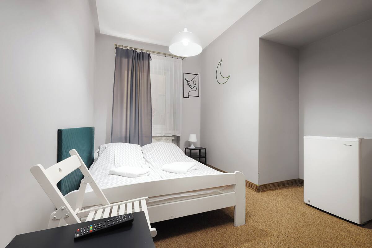Un dormitorio ordenado con una cama, un escritorio con una computadora portátil, un control remoto, una lámpara y un refrigerador.