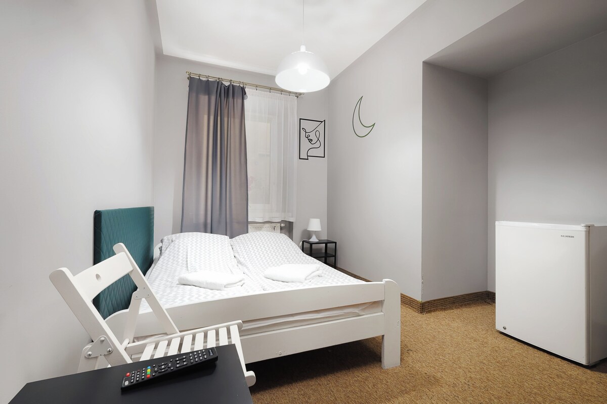 Een keurig opgeruimde slaapkamer met een bed, een bureau met een laptop, een afstandsbediening, een lamp en een koelkast.