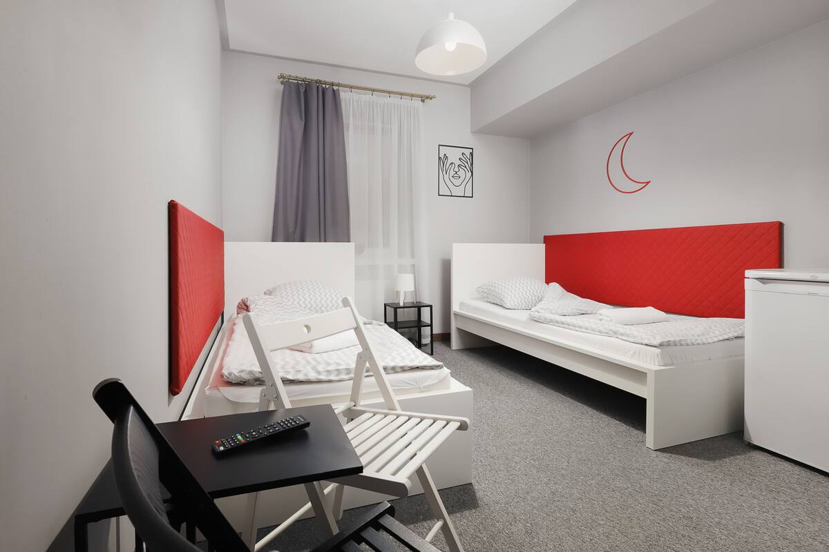 Una habitación de hotel moderna con una cama blanca, cabecera roja, un escritorio blanco con un control remoto y una silla gris.