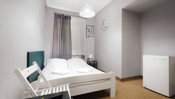 Un dormitorio ordenado con una cama, un escritorio con una computadora portátil, un control remoto, una lámpara y un refrigerador.