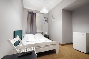 Ein Schlafzimmer mit Bett, Schreibtisch mit Laptop und Fernbedienung, kleiner Tisch mit Lampe, Kühlschrank und einem Fenster mit Vorhängen.