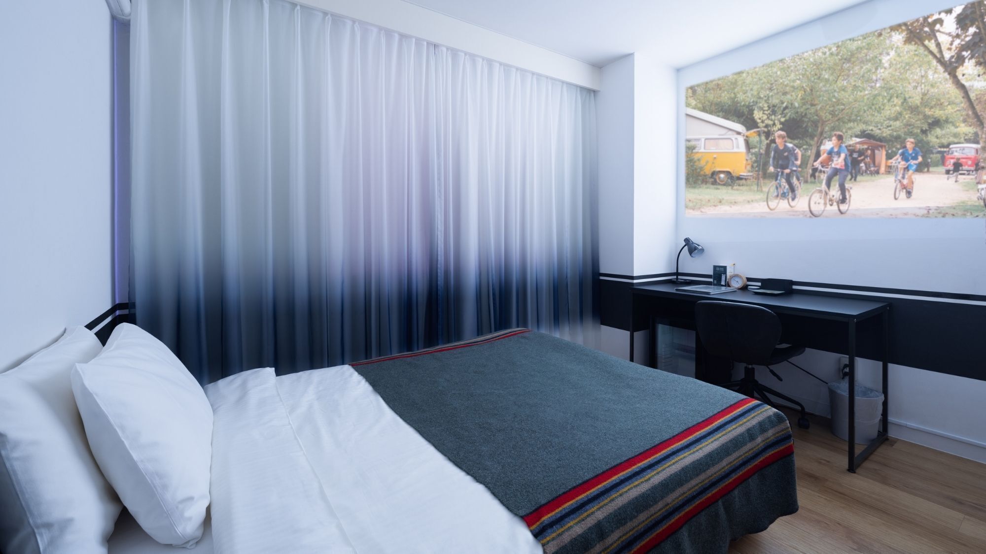 Habitació d'hotel amb un llit, una taula de treball i una finestra que mostra ciclistes.