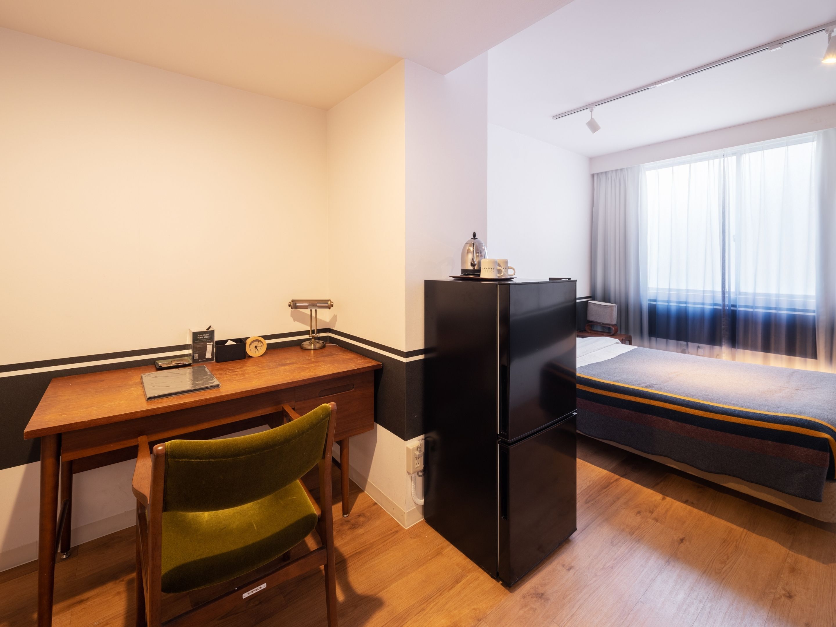 A compact hotel room with a wooden desk, a green chair, a black refrigerator, a bed with a striped blanket, and a window with curtains.