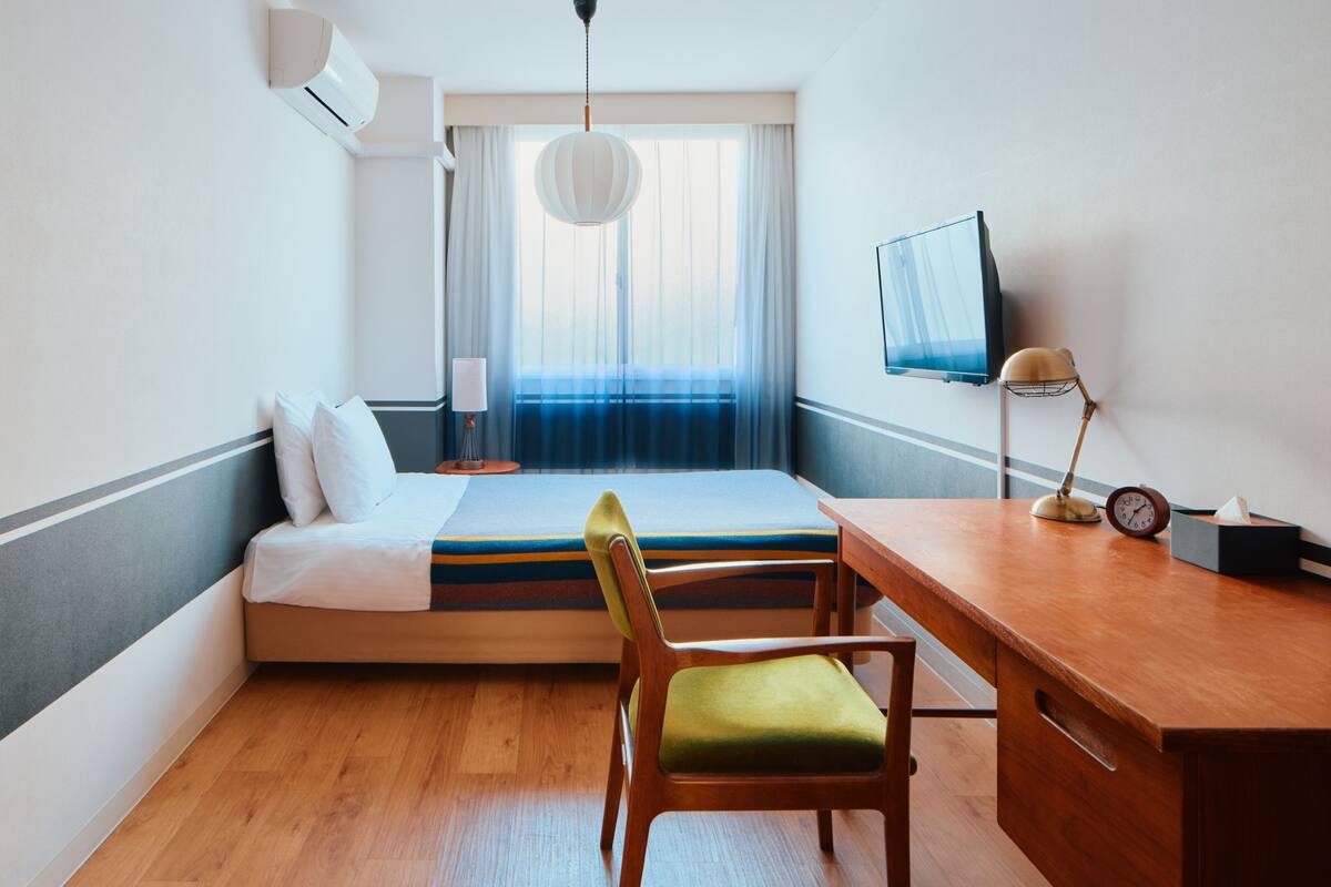 A hotel room with a bed, a desk, a chair, a TV, a lamp, and a clock.