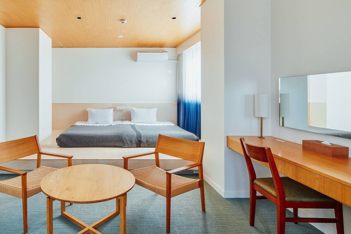 A hotel room with a bed, two wooden chairs, a small round table, a desk with a chair, a mirror, and a ceiling light fixture.
