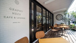 A cafe with a white brick wall, black-framed windows, and a large clock. Tables and chairs are outside.