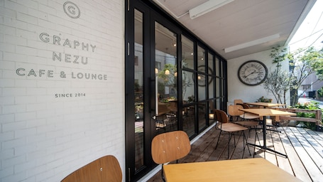 A cafe with a white brick wall, black-framed windows, and a large clock. Tables and chairs are outside.