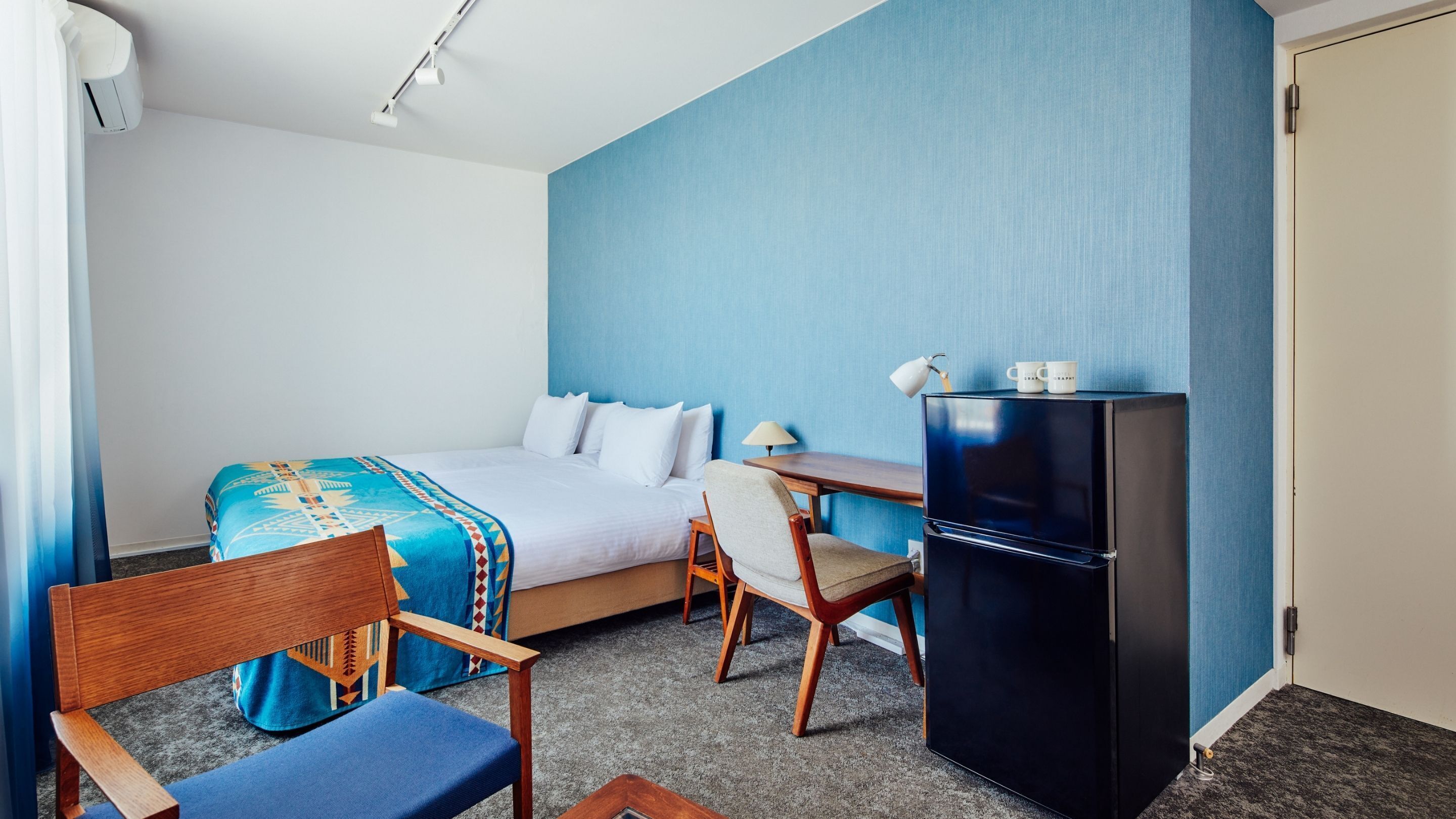 A hotel room with a bed, a desk, a chair, a refrigerator, and a lamp.