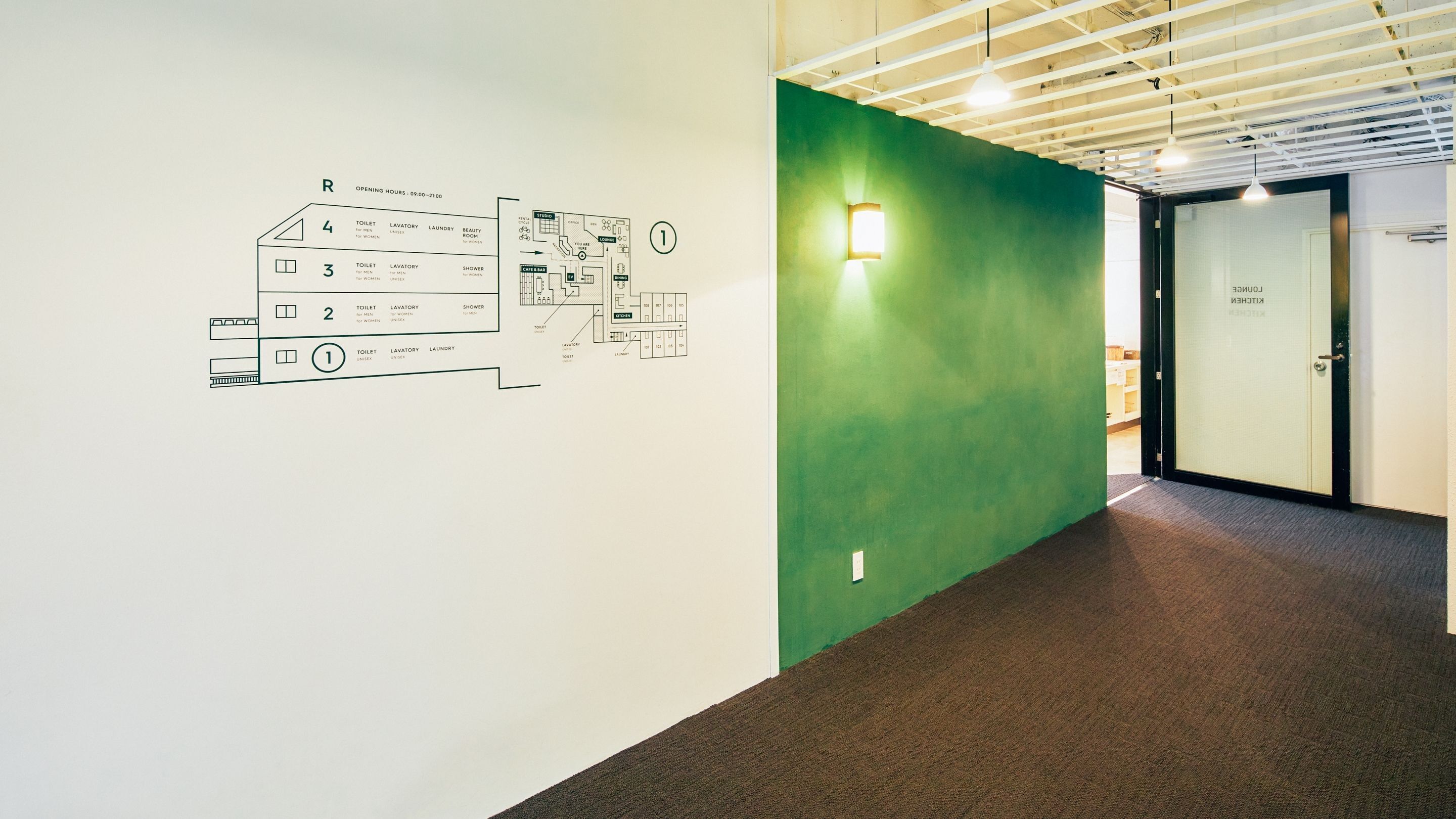 Un couloir d’hôtel avec un mur végétal, un plan des différents types de chambres et une porte portant l’inscription « Cuisine ».