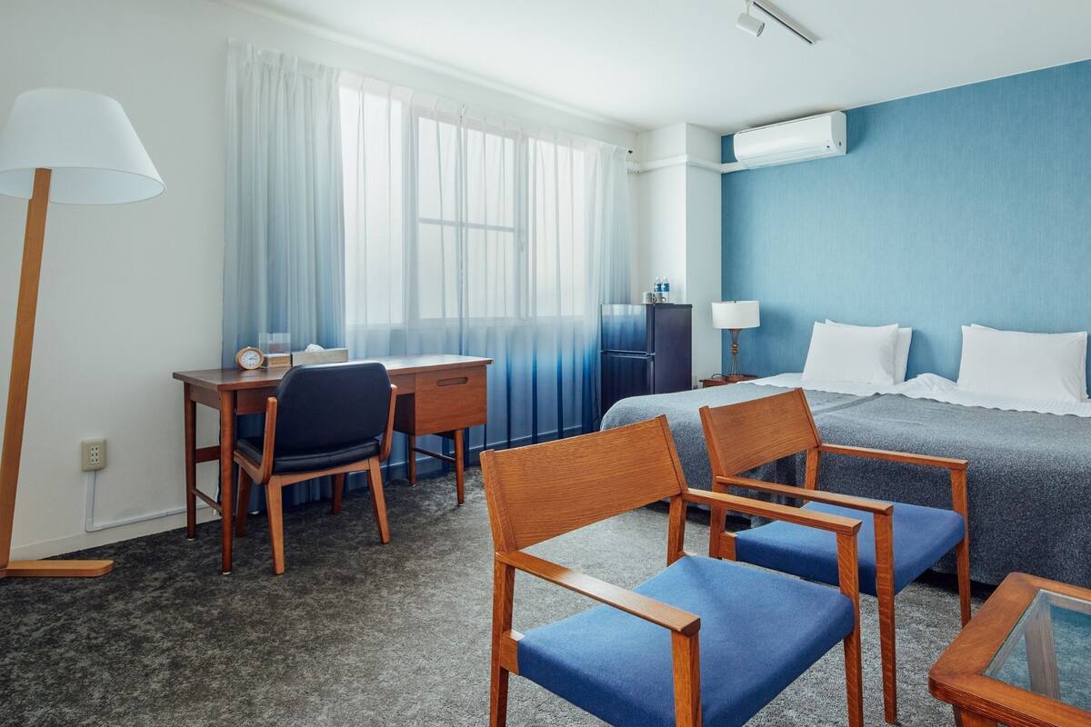 A hotel room with a bed, two chairs, a desk, a lamp, a TV, and a minibar.