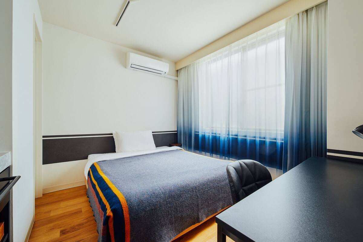 A hotel room with a bed, a desk, a chair, an air conditioning unit, and a window with sheer curtains.