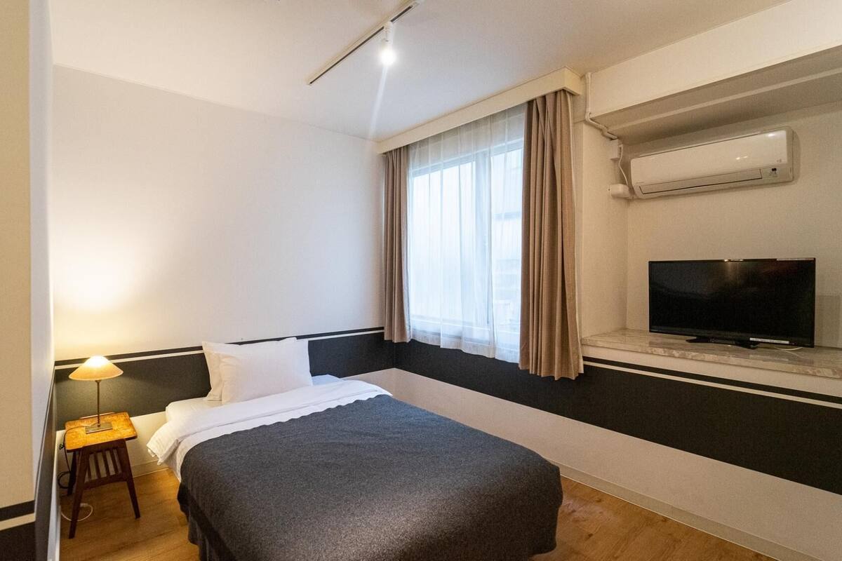 A hotel room with a bed, a TV, an air conditioning unit, and a window with curtains.