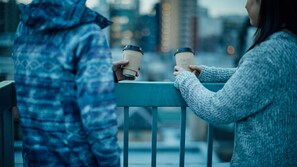 Two people holding coffee cups on a balcony with a cityscape in the background.