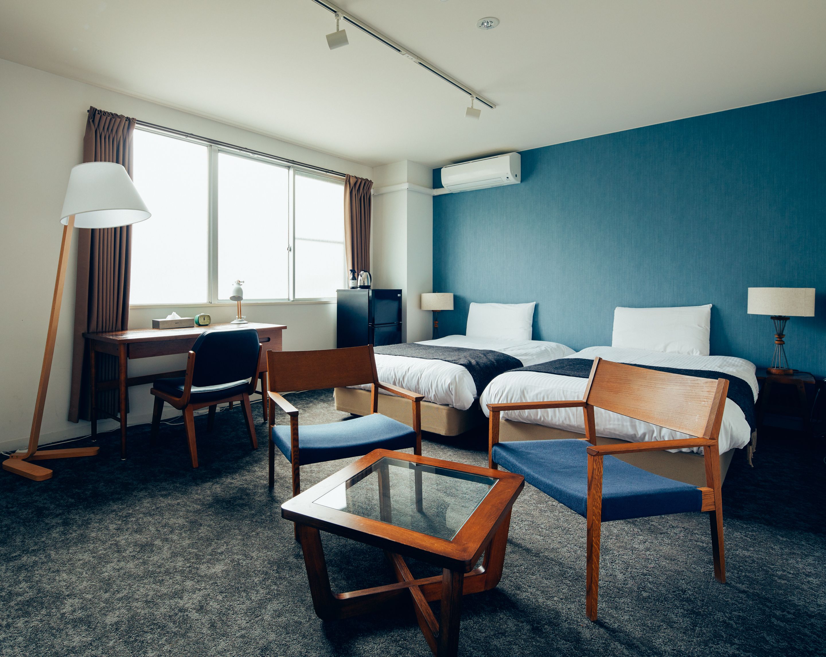 A hotel room with a desk, two beds, a chair, a table, and an air conditioning unit.