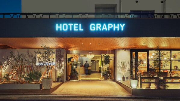 The exterior of Hotel Graphy with a prominent illuminated sign, a welcoming entrance, and potted plants around the entrance area.