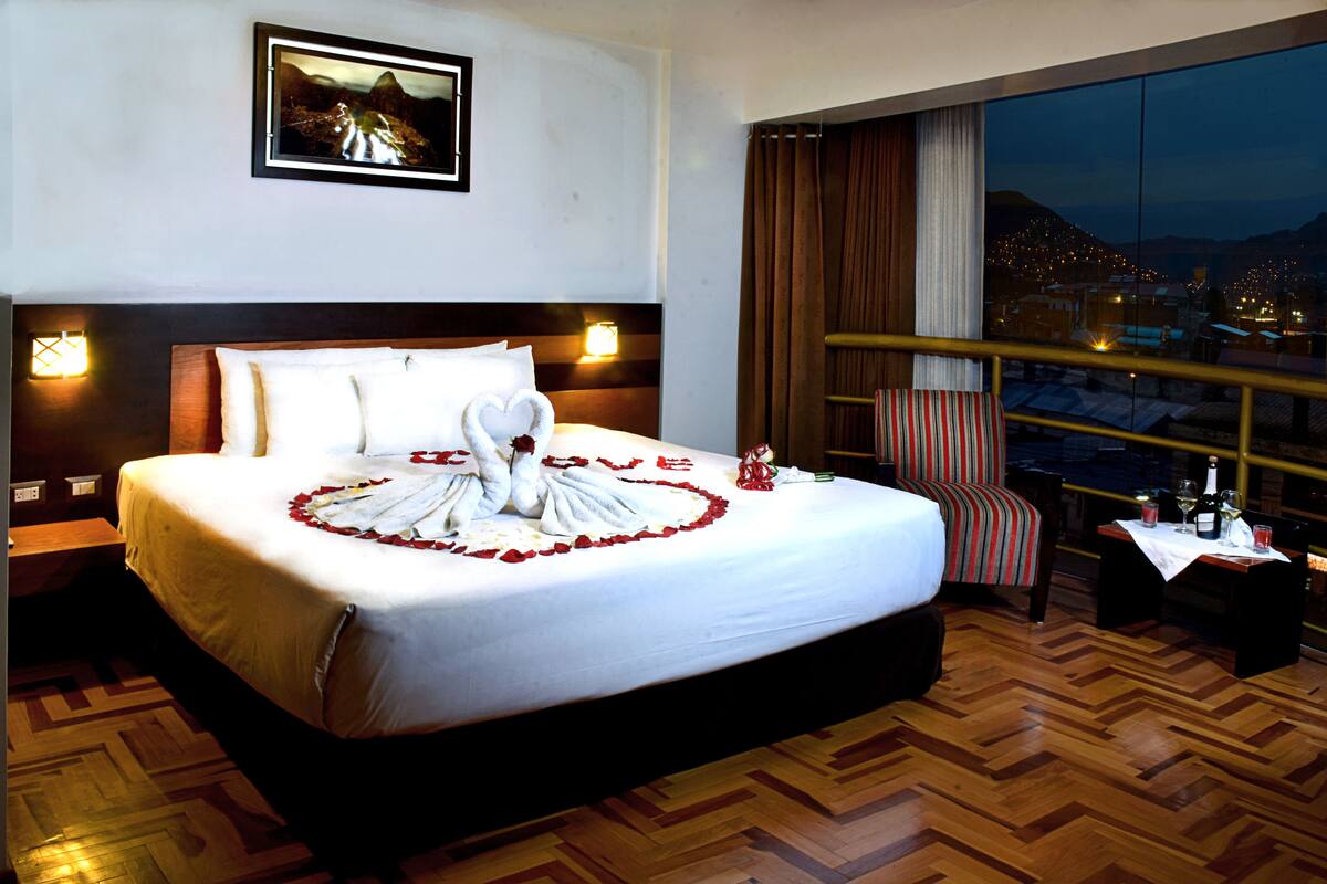 Ein Hotelzimmer mit einem großen Bett, Blick auf die nächtliche Stadt, einem gerahmten Bild an der Wand und einem Balkon mit Geländer.