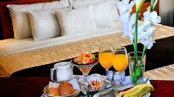 Une table préparée pour le petit-déjeuner, avec un lit en arrière-plan, un plateau contenant du pain, des viennoiseries et des fruits, et deux verres de jus d'orange.