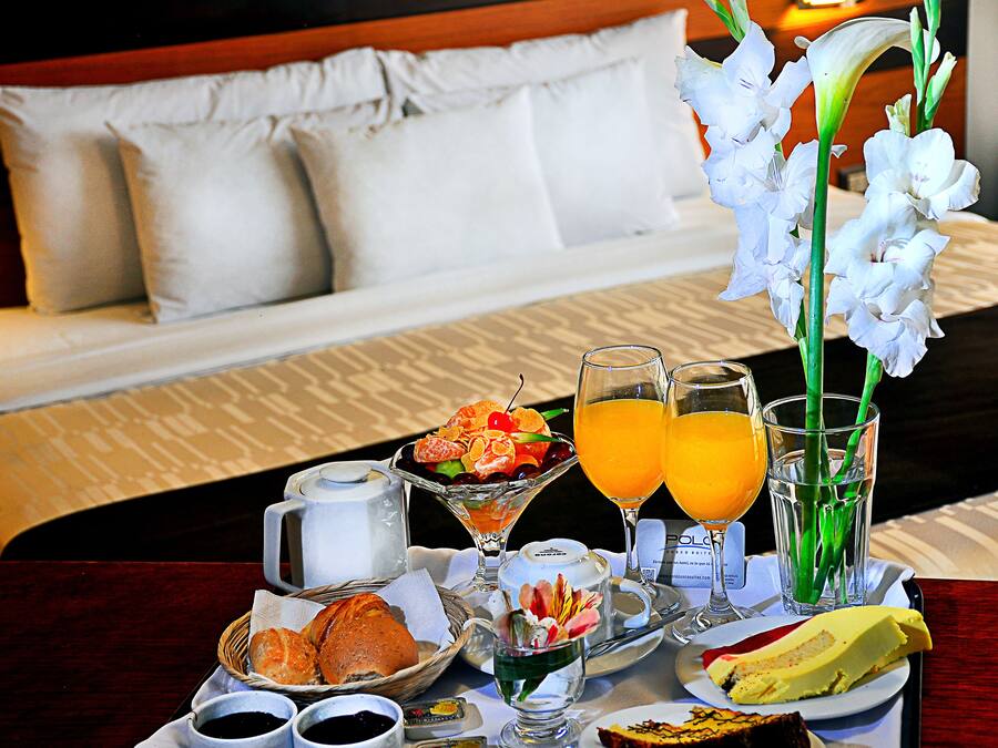 Colazione servita con un letto sullo sfondo, un vassoio con pane, pasticcini e frutta, e due bicchieri di succo d'arancia.
