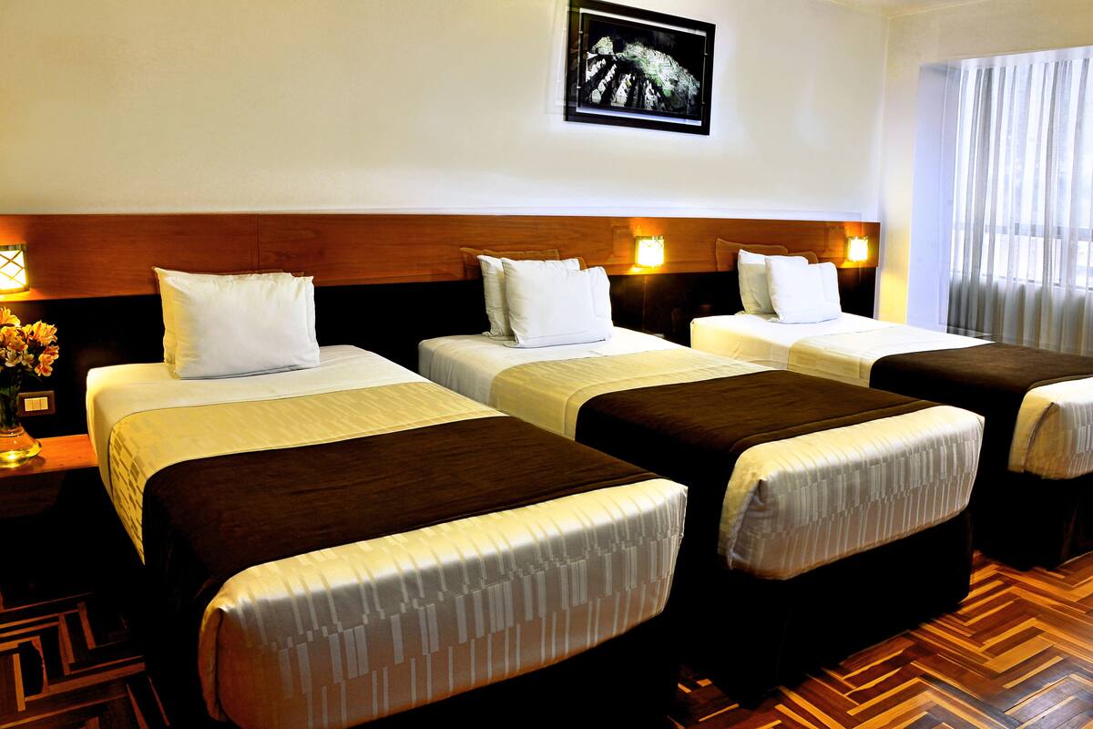 Ein Hotelzimmer mit vier nebeneinander stehenden Betten, einem hölzernen Kopfteil, einem gerahmten Bild an der Wand und einem gemusterten Boden.