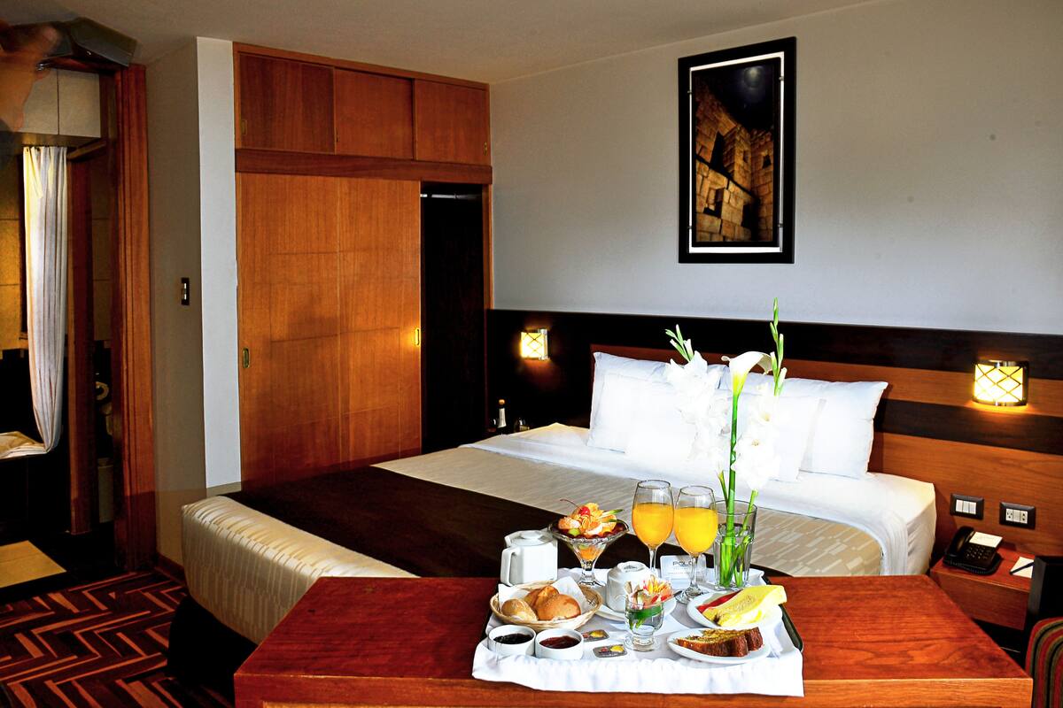Ein Hotelzimmer mit einem großen Bett, einem hölzernen Kopfteil, einem Nachttisch mit Lampe und einem Frühstücksbrett auf dem Tisch.
