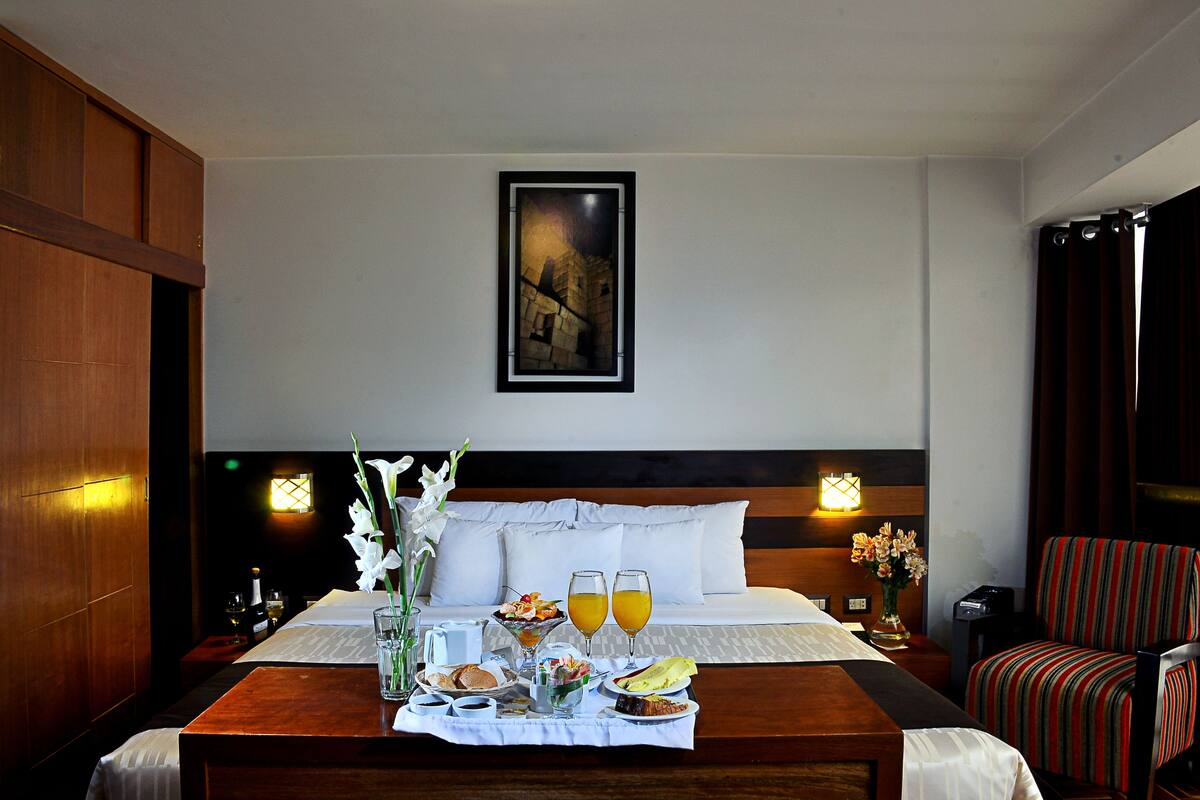 Ein Hotelzimmer mit Bett, Nachttischen, einem Esstisch mit Frühstück, einem Stuhl und einem Gemälde an der Wand.