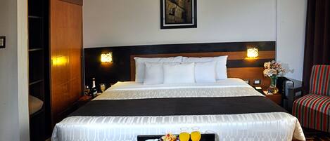 A hotel room with a large bed, a bedside table with a tray of breakfast, a nightstand with a lamp, and a framed picture on the wall.