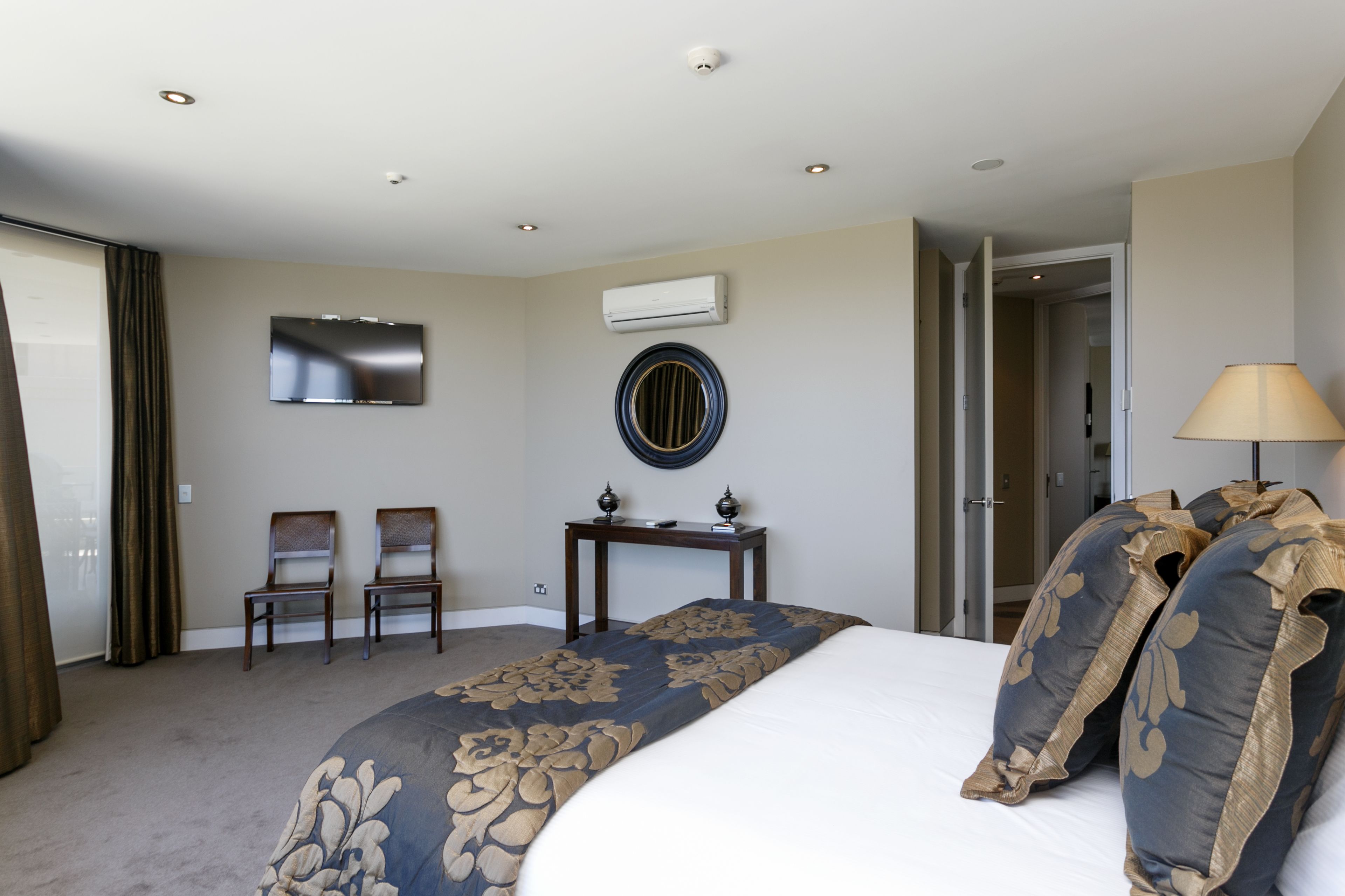A hotel room with a bed, two chairs, a small table, a TV, an air conditioning unit, and a lamp.