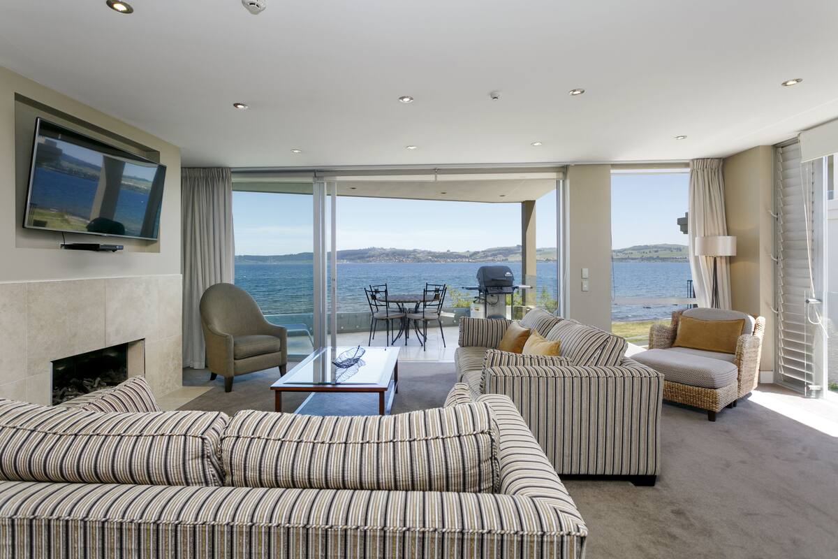 A spacious living room with a large TV, comfortable seating, a fireplace, and a view of the sea.