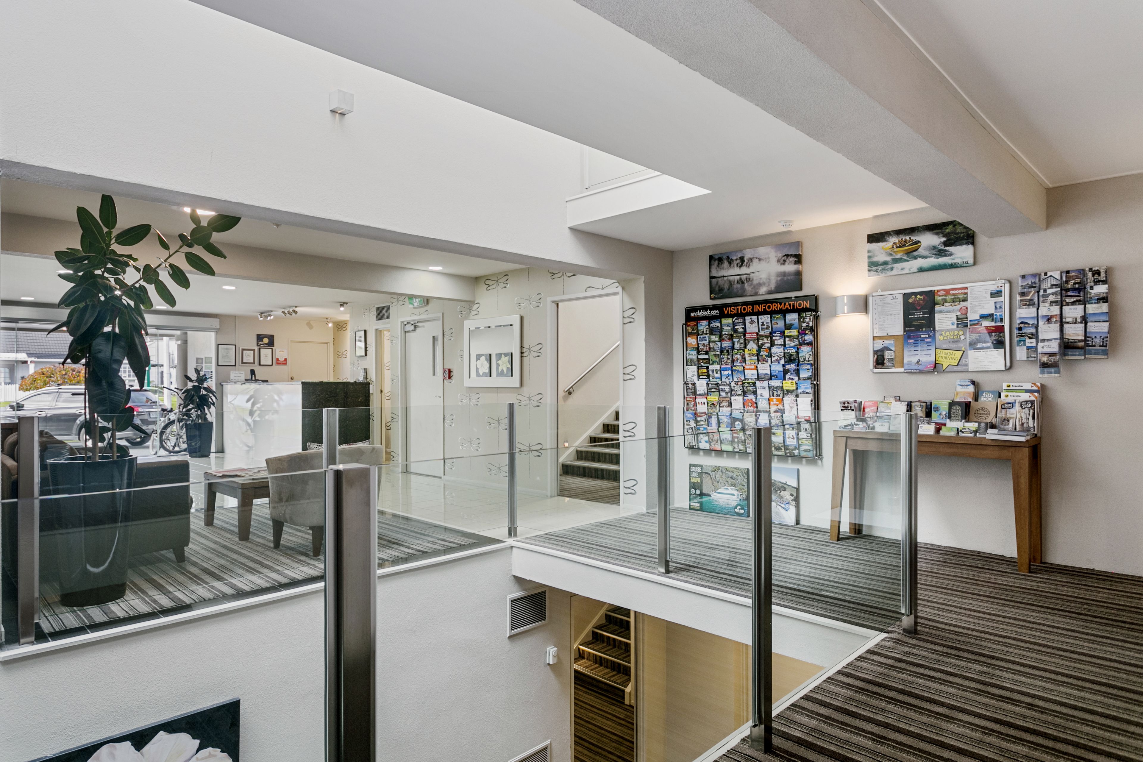 A modern hotel lobby with a glass railing, a staircase, and a wall displaying various brochures and a 'Visitor Information' sign.