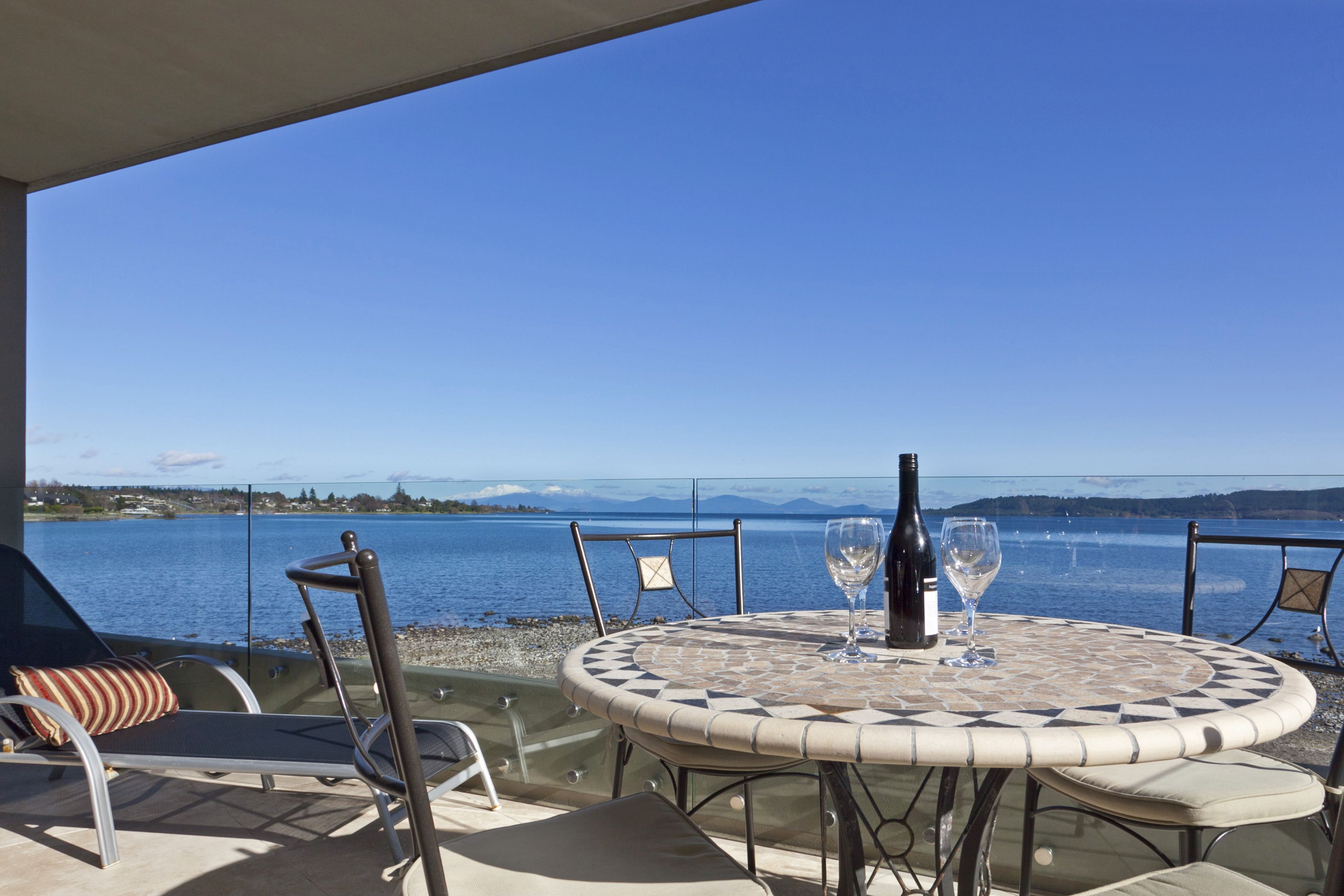 A round table with a mosaic pattern, two wine glasses, and a bottle of wine on a terrace overlooking a lake with mountains in the distance.