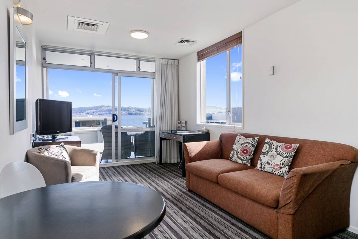 A modern hotel room with a large window, a sofa, a dining table, and a TV.