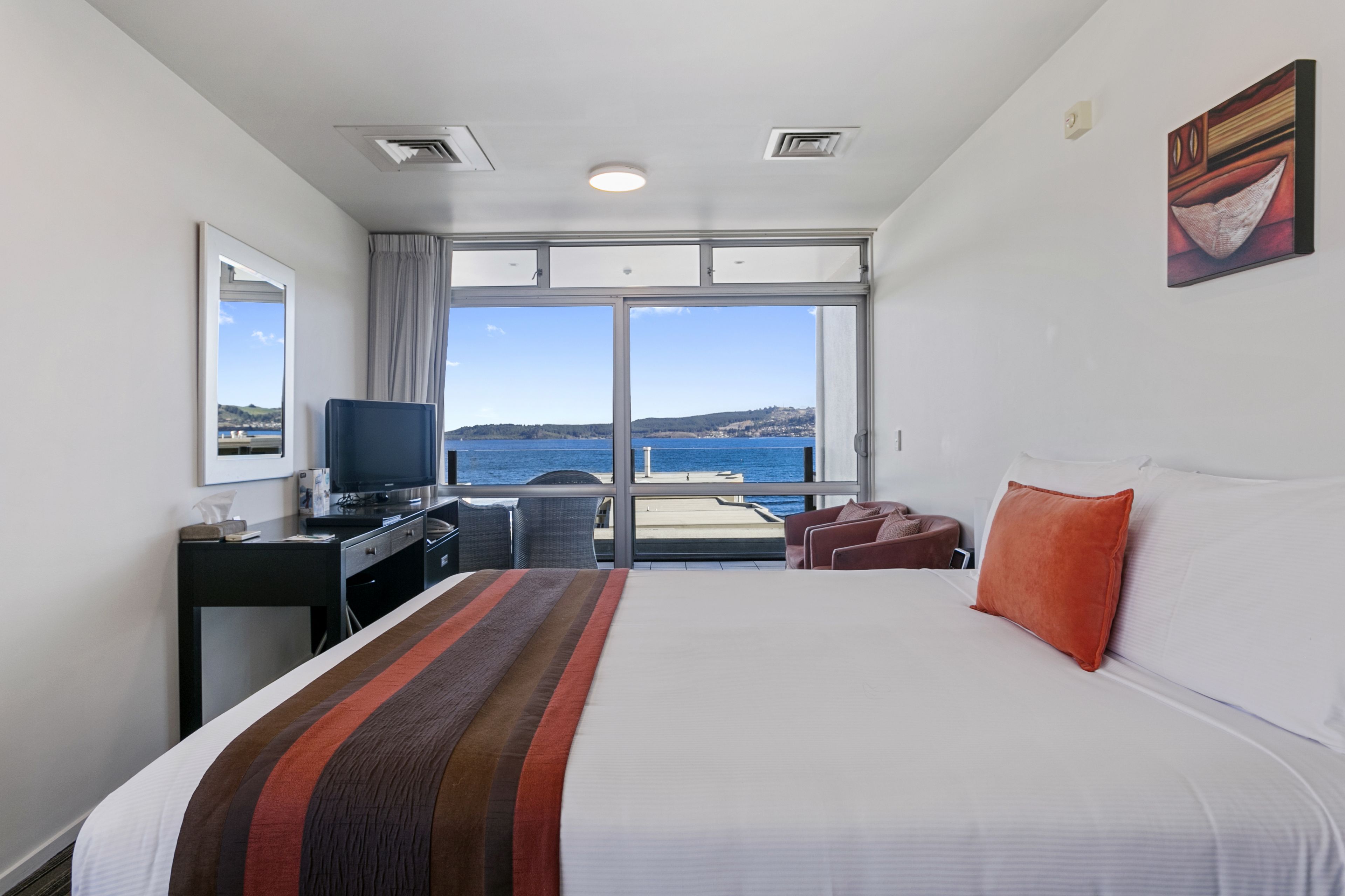 Habitación de hotel con una cama grande, un escritorio con ordenador, televisión y vistas al mar a través de amplios ventanales.