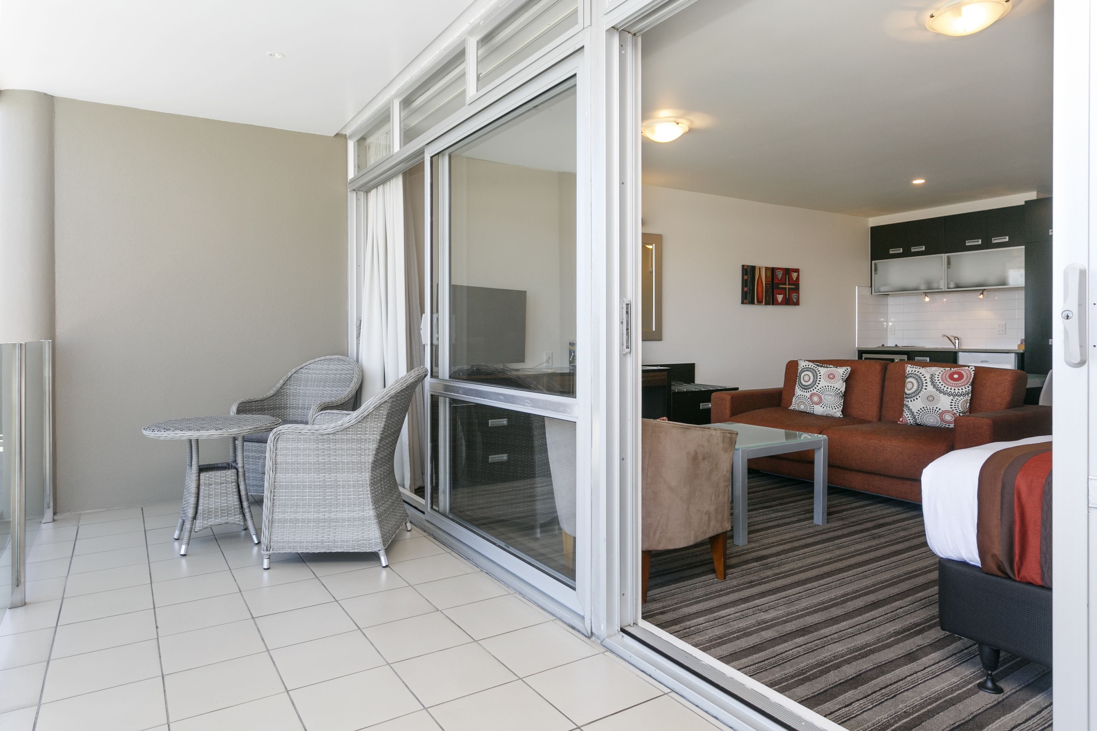 A modern living room with a glass door leading to a balcony, a sofa, a small table, and a kitchen area visible through the glass.
