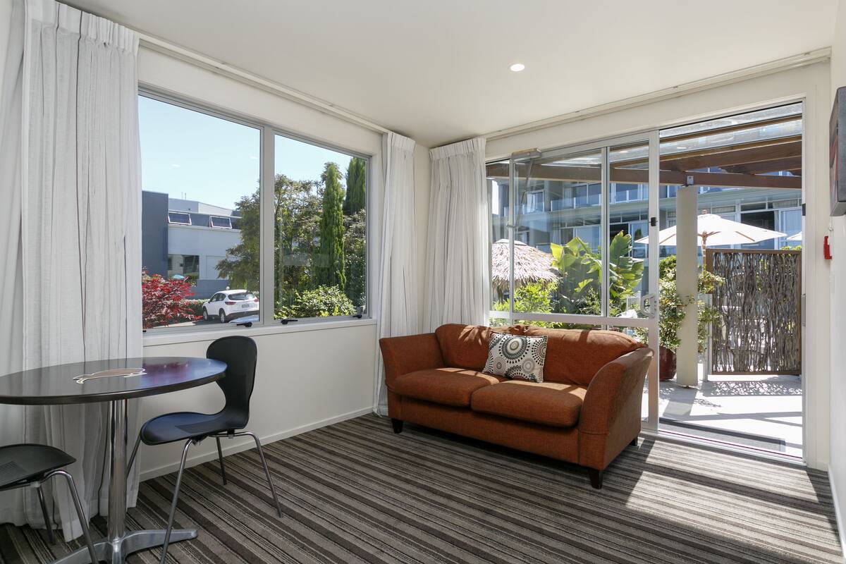 A modern living room with a sofa, a small table, and chairs. There is a large window with curtains, a sliding glass door leading to an outdoor area with umbrellas.