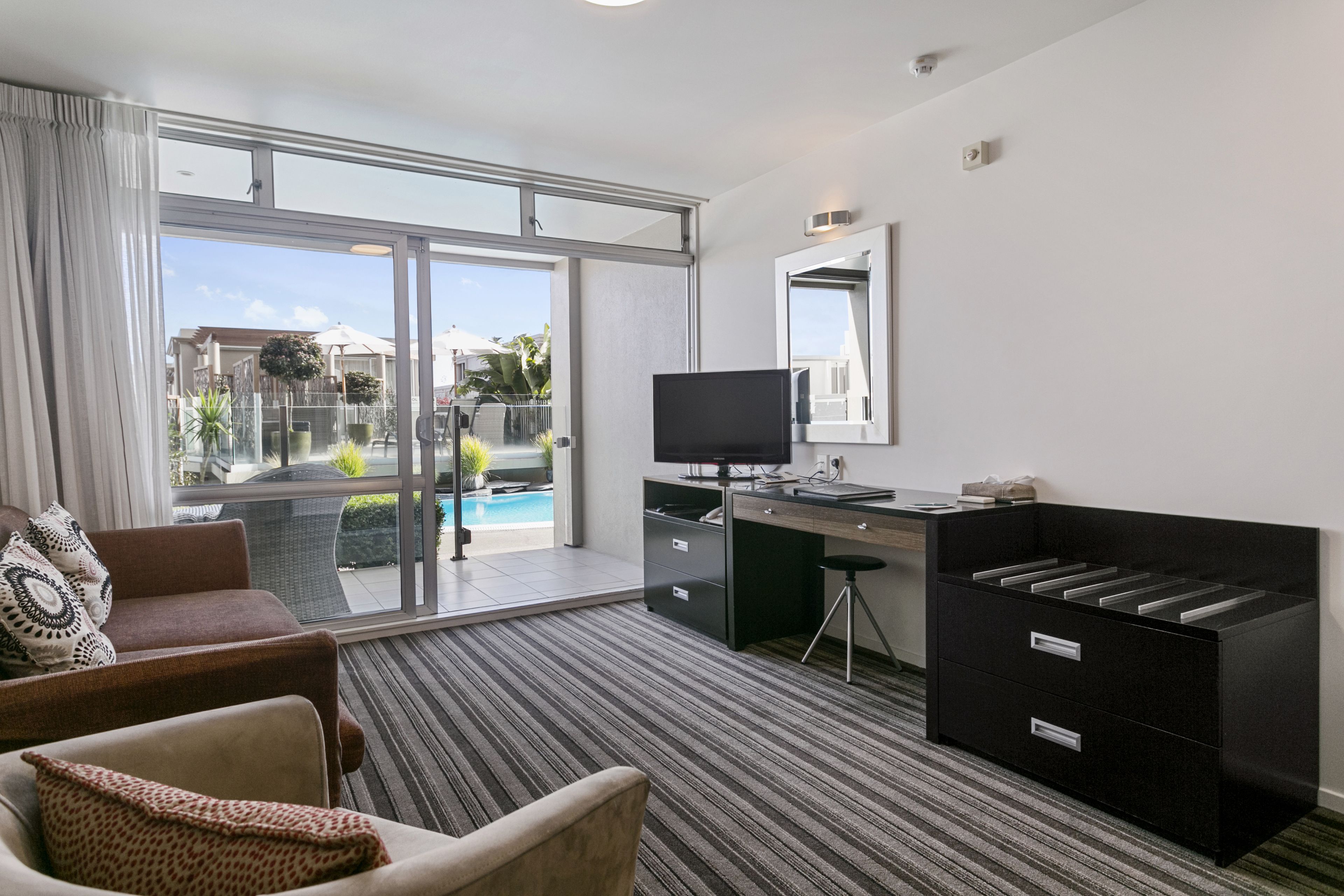 A hotel room with a large sliding glass door leading to a balcony, a desk with a flat-screen TV, a comfortable sofa, and a black filing cabinet.