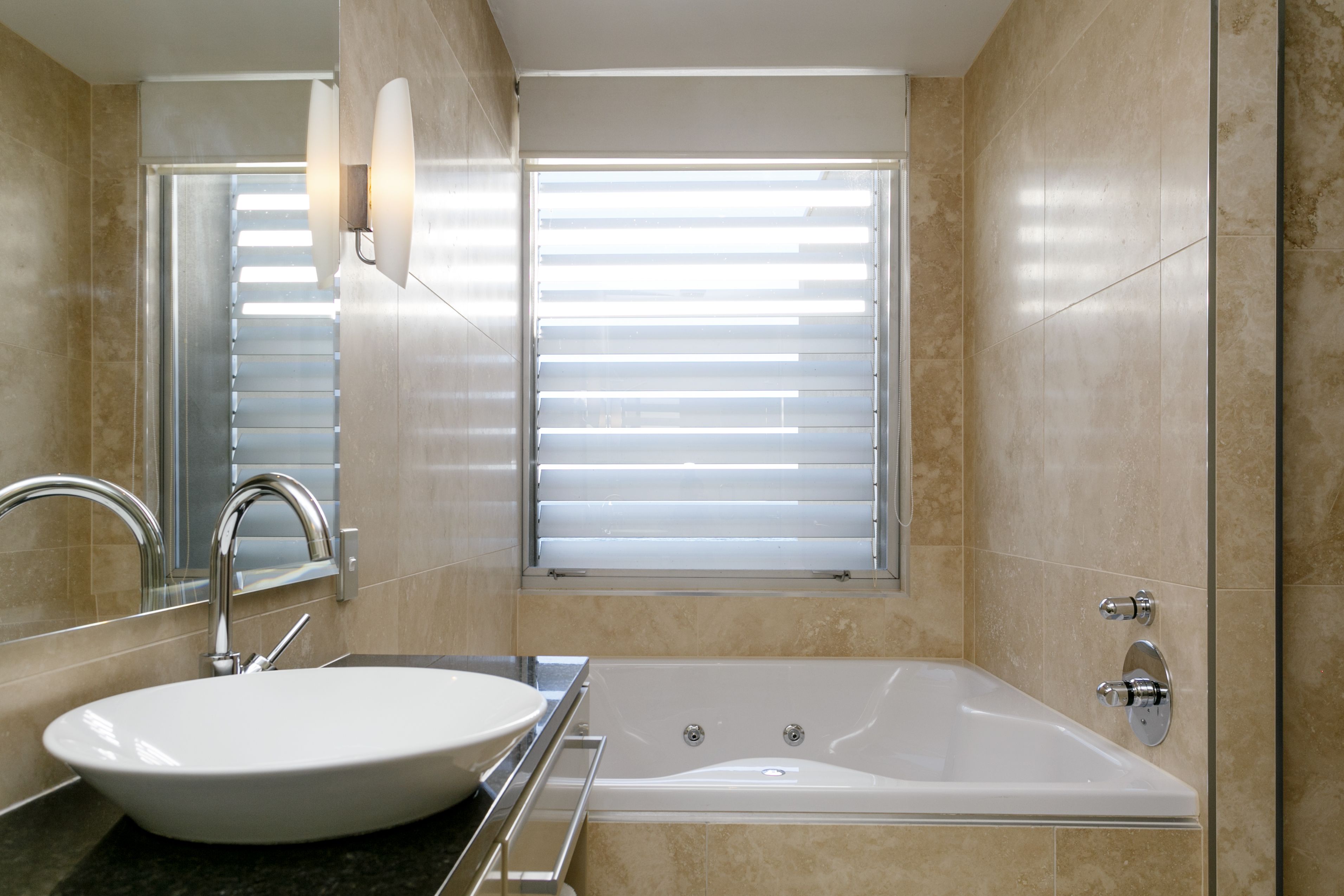 Un baño moderno con un lavamanos ovalado grande, una bañera independiente y una ventana con persianas.