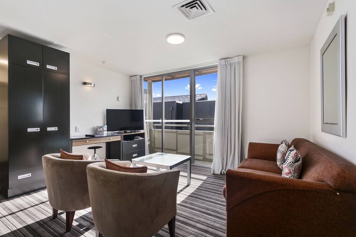 A modern hotel room with a large window, a flat-screen TV, a sofa, and a dining area with a table and chairs.
