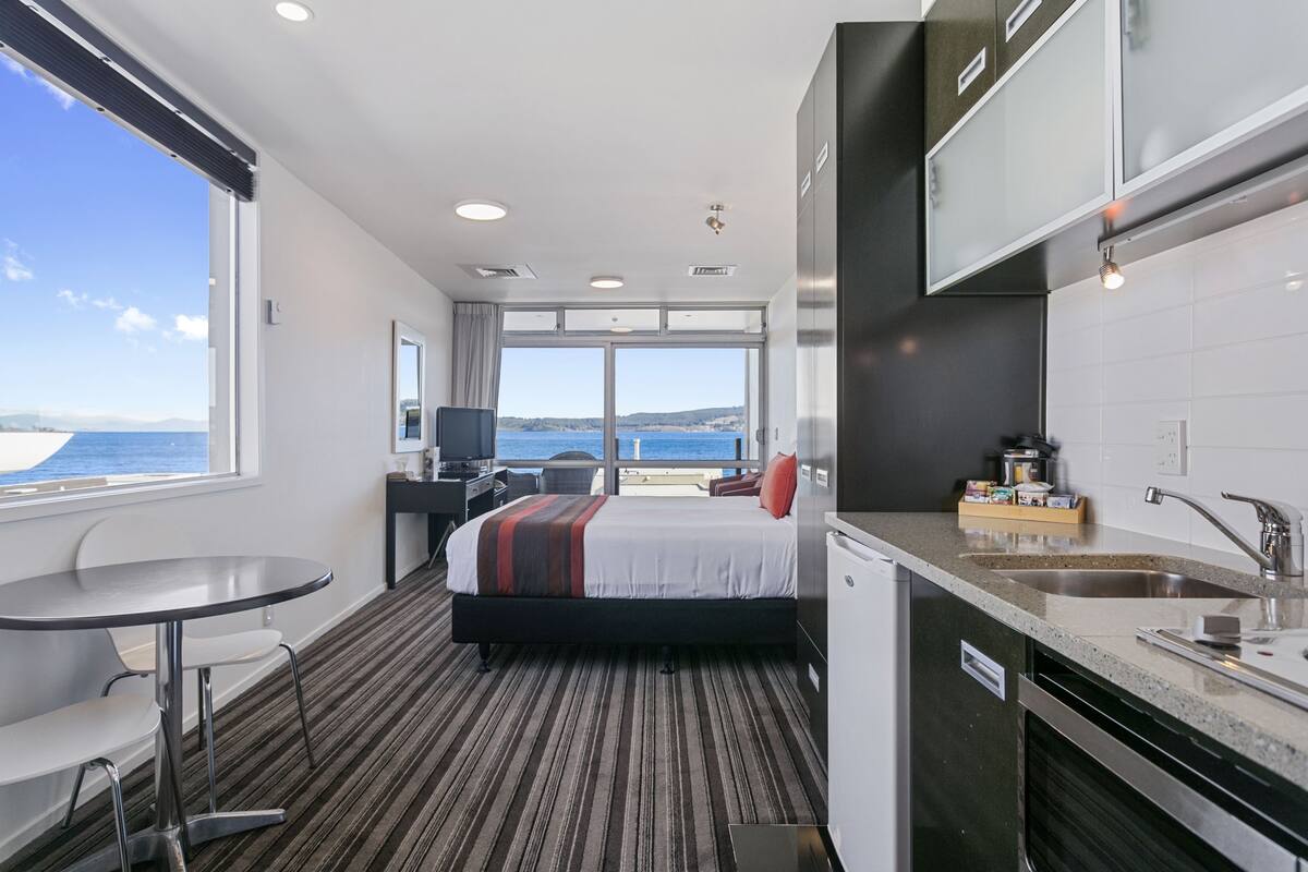 A hotel room with a bed, a desk, a chair, a small table, a sink, and a view of the sea.
