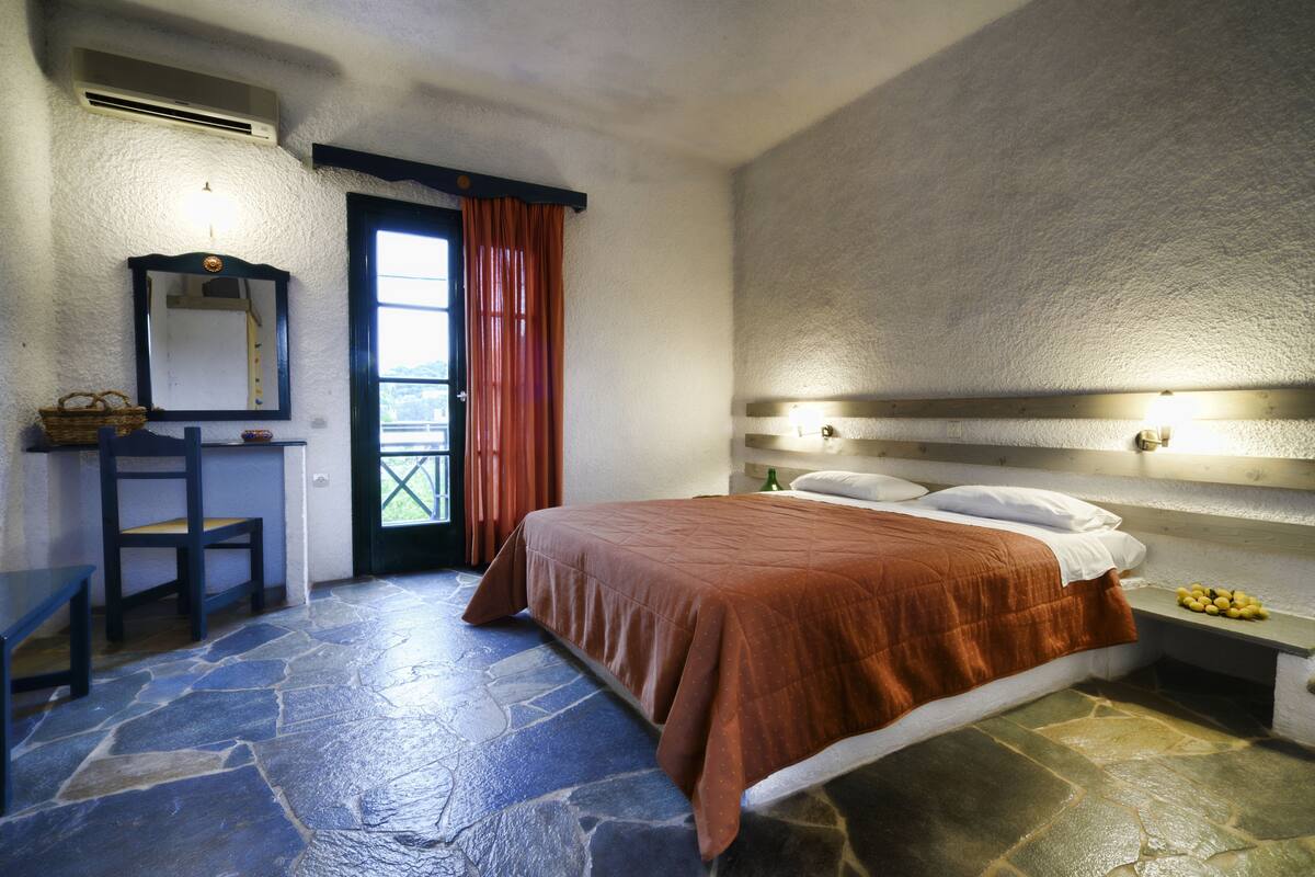 A bedroom with a stone floor, a bed with a brown bedspread, a nightstand with a lamp, a mirror, and a window with curtains.