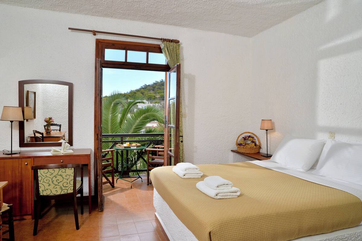 A hotel room with a bed, a desk, a chair, a small table, and a balcony with a view of palm trees.